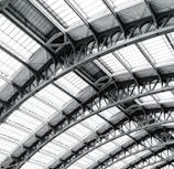 Intricate architectural structure featuring a series of curved metal and glass panels that form a dome-like pattern. The design showcases engineering complexity with symmetrical and repetitive elements creating a visually captivating ceiling.