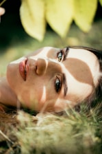 A person is lying on the grass, with sunlight filtering through leaves casting shadows on their face. Their eyes are open, and they are gazing upwards. The scene conveys a sense of tranquility and connection with nature.