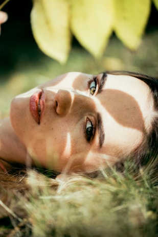 A person is lying on the grass, with sunlight filtering through leaves casting shadows on their face. Their eyes are open, and they are gazing upwards. The scene conveys a sense of tranquility and connection with nature.