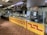 A well-lit interior of a traditional Mexican restaurant with a display counter filled with various food items. The counter is adorned with yellow and red tiles in a geometric pattern. A sign labeled 'Menudo' is visible above the counter. Festive decorations in green, white, and red hang from the ceiling, conveying a cultural or festive theme. The kitchen area and various kitchen equipment are seen behind the counter, and a tiled floor completes the look.