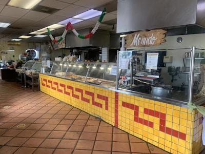 A well-lit interior of a traditional Mexican restaurant with a display counter filled with various food items. The counter is adorned with yellow and red tiles in a geometric pattern. A sign labeled 'Menudo' is visible above the counter. Festive decorations in green, white, and red hang from the ceiling, conveying a cultural or festive theme. The kitchen area and various kitchen equipment are seen behind the counter, and a tiled floor completes the look.