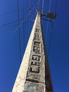 A tall concrete utility pole rises upwards against a vibrant blue sky. It is embedded with numbers or characters and is connected to multiple wires extending in various directions. A utility box or panel is mounted near the top.