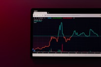 A tablet screen displaying a financial chart with two lines, one red and one cyan, representing data trends over time. The background is dark with text showing figures at the top, including maximum, minimum values, and a percentage.