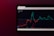A tablet screen displaying a financial chart with two lines, one red and one cyan, representing data trends over time. The background is dark with text showing figures at the top, including maximum, minimum values, and a percentage.
