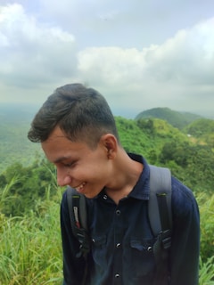 A young person with short hair is smiling while standing outdoors. They are wearing a dark blue shirt and carrying a backpack. The background features lush green hills and cloudy skies, indicating a natural, mountainous setting.