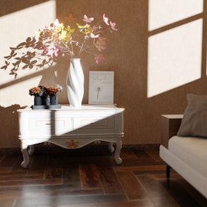 A stylish, sunlit interior features a white ornate table against a beige wall. On the table, a white vase with delicate, colorful leaves casts intricate shadows. Beside it is a book titled 'Learning to Breathe Again' and a set of three small, dark vases with dried flowers. The wooden floor and light-colored couch contribute to the room's warm aesthetic.