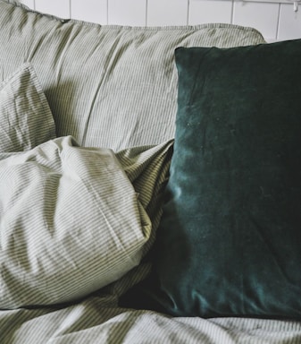A cozy arrangement featuring a white and green pinstriped cushion, accompanied by a solid dark green velvet pillow. The setting appears to be on a plush seating or bedding area in a softly lit room.
