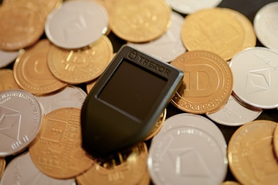 A mix of various cryptocurrency coins including Bitcoin and Dogecoin piled together. In the center lies a black Trezor hardware wallet, used for storing cryptocurrencies securely.