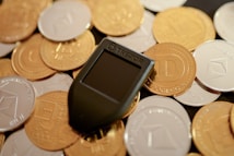 A mix of various cryptocurrency coins including Bitcoin and Dogecoin piled together. In the center lies a black Trezor hardware wallet, used for storing cryptocurrencies securely.