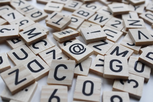 Wooden tiles with black letters and symbols are scattered across a surface. The tiles resemble those used in word games, featuring a variety of letters, numbers, and punctuation marks such as '@' and '#'.