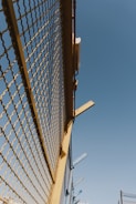 A metal fence with yellow framing and a mesh design stretches upward against a clear blue sky. There are some metal beams protruding from the top of the fence at angles.