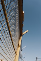 A metal fence with yellow framing and a mesh design stretches upward against a clear blue sky. There are some metal beams protruding from the top of the fence at angles.