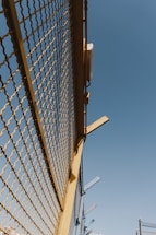 A metal fence with yellow framing and a mesh design stretches upward against a clear blue sky. There are some metal beams protruding from the top of the fence at angles.