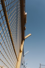 A metal fence with yellow framing and a mesh design stretches upward against a clear blue sky. There are some metal beams protruding from the top of the fence at angles.
