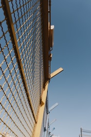 A metal fence with yellow framing and a mesh design stretches upward against a clear blue sky. There are some metal beams protruding from the top of the fence at angles.