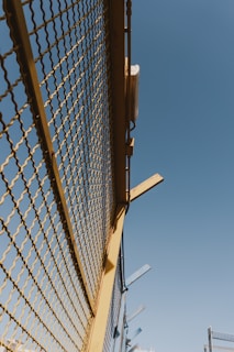 A metal fence with yellow framing and a mesh design stretches upward against a clear blue sky. There are some metal beams protruding from the top of the fence at angles.