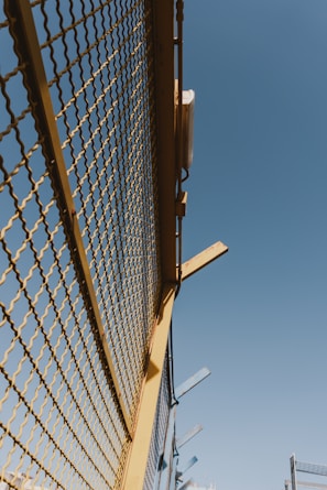 A metal fence with yellow framing and a mesh design stretches upward against a clear blue sky. There are some metal beams protruding from the top of the fence at angles.