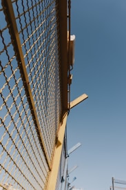 A metal fence with yellow framing and a mesh design stretches upward against a clear blue sky. There are some metal beams protruding from the top of the fence at angles.