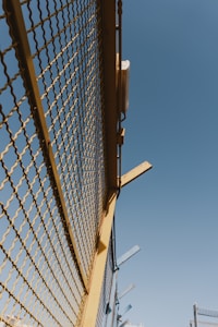 A metal fence with yellow framing and a mesh design stretches upward against a clear blue sky. There are some metal beams protruding from the top of the fence at angles.