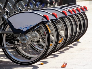 A line of bicycles neatly parked in a bike-sharing station. The bicycles have a modern design with white protective covers above the wheels and distinct red elements on the back. A few autumn leaves are scattered on the ground, indicating a crisp fall day.