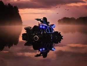An off-road vehicle with bright blue lights is partially submerged in water, reflecting the dramatic colors of the sunset sky. The surrounding scenery includes silhouettes of trees and birds flying in the background, creating a misty and mysterious atmosphere.