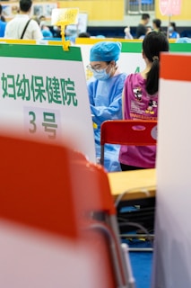 A healthcare professional dressed in blue scrubs, a cap, and a mask is seated next to a person wearing a pink shirt with cartoon characters on the back. They are in what appears to be a clinical setting with various signs and partitions around them.