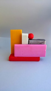 A playful arrangement of colorful geometric shapes including rectangular blocks in yellow, pink, white, gray, and red, along with a small red sphere placed on top. The scene is set against a neutral gray background, focusing on the vivid colors and simple forms of the objects.