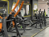 A gym with various workout machines including weightlifting equipment like benches and racks. The room has large mirrors on the walls, orange and black exercise machines, and visible air conditioning units. The flooring is padded, designed for safety and comfort during physical exercise.