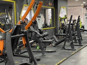A gym with various workout machines including weightlifting equipment like benches and racks. The room has large mirrors on the walls, orange and black exercise machines, and visible air conditioning units. The flooring is padded, designed for safety and comfort during physical exercise.