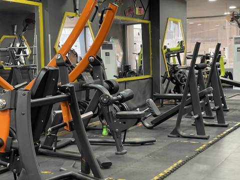 A gym with various workout machines including weightlifting equipment like benches and racks. The room has large mirrors on the walls, orange and black exercise machines, and visible air conditioning units. The flooring is padded, designed for safety and comfort during physical exercise.