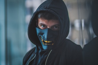 A person wears a black hooded sweatshirt and a dark mask with a bold, stylized facial design. The mask covers the lower half of the face, and the eyes are visible, giving a mysterious and enigmatic look. The background is blurred, focusing attention on the person.