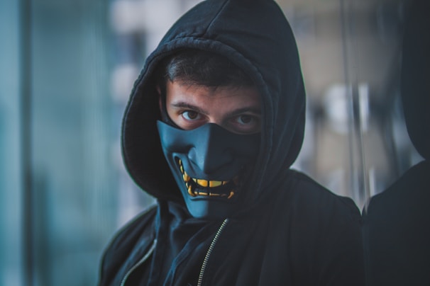 A person wears a black hooded sweatshirt and a dark mask with a bold, stylized facial design. The mask covers the lower half of the face, and the eyes are visible, giving a mysterious and enigmatic look. The background is blurred, focusing attention on the person.
