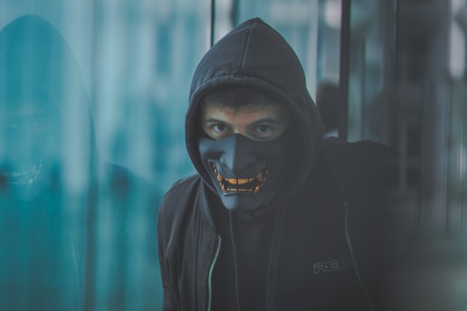 A person wearing a dark hoodie and a mask with orange accents stands in a mysterious setting. The reflection on a glass surface adds depth and intrigue.
