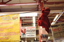 A market setting with various advertisements for poultry and seafood. Hanging prominently is a large red lobster plush toy. Surrounding signs promote locally sourced poultry and seafood from Ohio. The overhead structure is made of red metal beams, creating an industrial feel.