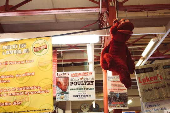 A market setting with various advertisements for poultry and seafood. Hanging prominently is a large red lobster plush toy. Surrounding signs promote locally sourced poultry and seafood from Ohio. The overhead structure is made of red metal beams, creating an industrial feel.