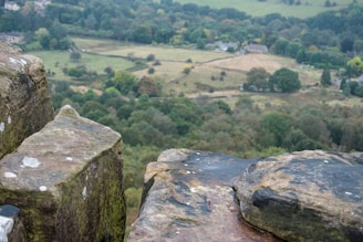 Weathered sandstone rocks are in the foreground with a view overlooking a lush, green landscape featuring rolling hills, patches of trees, and scattered houses. The scene is peaceful and expansive, capturing a sense of natural beauty and tranquility.