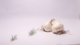 Two heads of garlic are placed on a white surface with their roots visible. There are also small green herbs scattered around them.