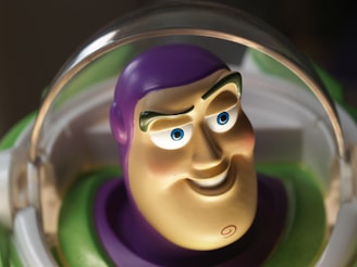 A close-up image of a toy character with a large, smiling face. The character has a purple helmet or head covering and green and white outerwear. The toy's facial features include large blue eyes, raised eyebrows, and a pronounced chin with a distinctive swirl symbol.