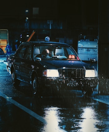 A taxi moving through a wet, dimly lit urban environment at night. The reflection of streetlights and surrounding elements is visible in the wet pavement. Two people are inside the vehicle, with one seemingly looking at a device.