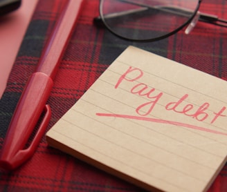 a note that says pay debt next to a pen and glasses