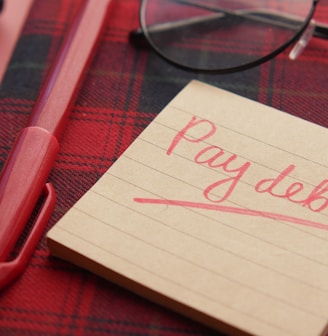 a note that says pay debt next to a pen and glasses