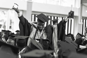 A jubilant graduate wearing a cap and gown is raising his arms triumphantly, holding a diploma and a smartphone. He is surrounded by other graduates who are also wearing caps and gowns, creating a festive and celebratory atmosphere.