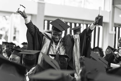 A jubilant graduate wearing a cap and gown is raising his arms triumphantly, holding a diploma and a smartphone. He is surrounded by other graduates who are also wearing caps and gowns, creating a festive and celebratory atmosphere.