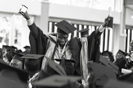 A jubilant graduate wearing a cap and gown is raising his arms triumphantly, holding a diploma and a smartphone. He is surrounded by other graduates who are also wearing caps and gowns, creating a festive and celebratory atmosphere.
