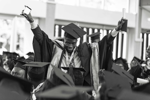 A jubilant graduate wearing a cap and gown is raising his arms triumphantly, holding a diploma and a smartphone. He is surrounded by other graduates who are also wearing caps and gowns, creating a festive and celebratory atmosphere.