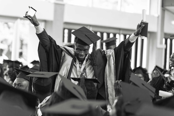 A jubilant graduate wearing a cap and gown is raising his arms triumphantly, holding a diploma and a smartphone. He is surrounded by other graduates who are also wearing caps and gowns, creating a festive and celebratory atmosphere.
