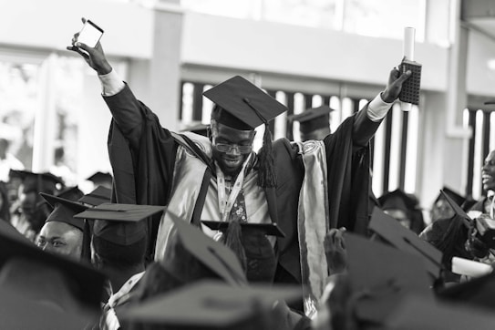 A jubilant graduate wearing a cap and gown is raising his arms triumphantly, holding a diploma and a smartphone. He is surrounded by other graduates who are also wearing caps and gowns, creating a festive and celebratory atmosphere.