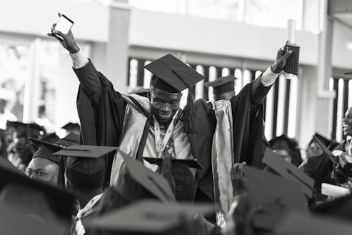 A jubilant graduate wearing a cap and gown is raising his arms triumphantly, holding a diploma and a smartphone. He is surrounded by other graduates who are also wearing caps and gowns, creating a festive and celebratory atmosphere.