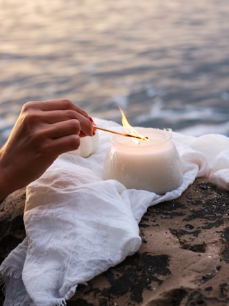 A hand is seen lighting a white candle with a match on a rocky surface near water. The scene is serene, with a white cloth draped under the candle. The flames from the match and candle create a warm, gentle glow amidst the soft, ambient light of the setting.