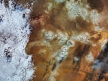An aerial view of a geothermal landscape with steam vents and patches of white mineral deposits. The area is predominantly brown with various shades, interspersed with white and blue on one side, indicative of snow or frost. There are several circular formations suggesting volcanic activity.